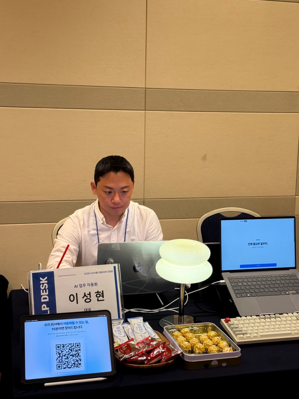 2026 수산식품기업 바우처 간담회 상담 부스에서 노트북으로 일하는 이성현. 책상 위 태블릿에 15분 AI 진단 QR 안내가 있다.